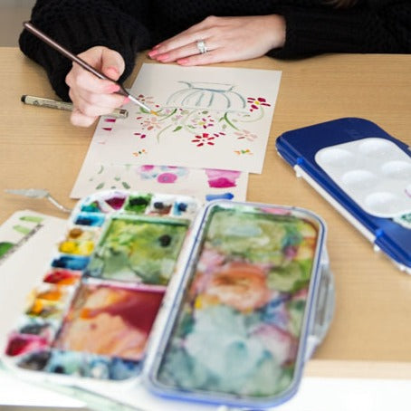 A pair of hands painting a floral design on paper using watercolor paints