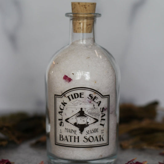 Small glass jar with cork stopper, filled with bath soaking salts and surrounded by rose petals